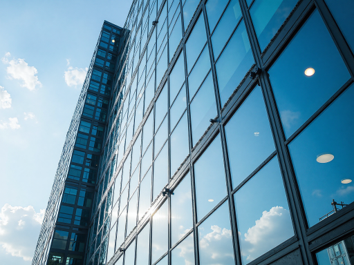 Modern office building exterior with glass facade, reflecting blue sky and white clouds under sunlight, surrounded by green plants, showcasing modern business environment
